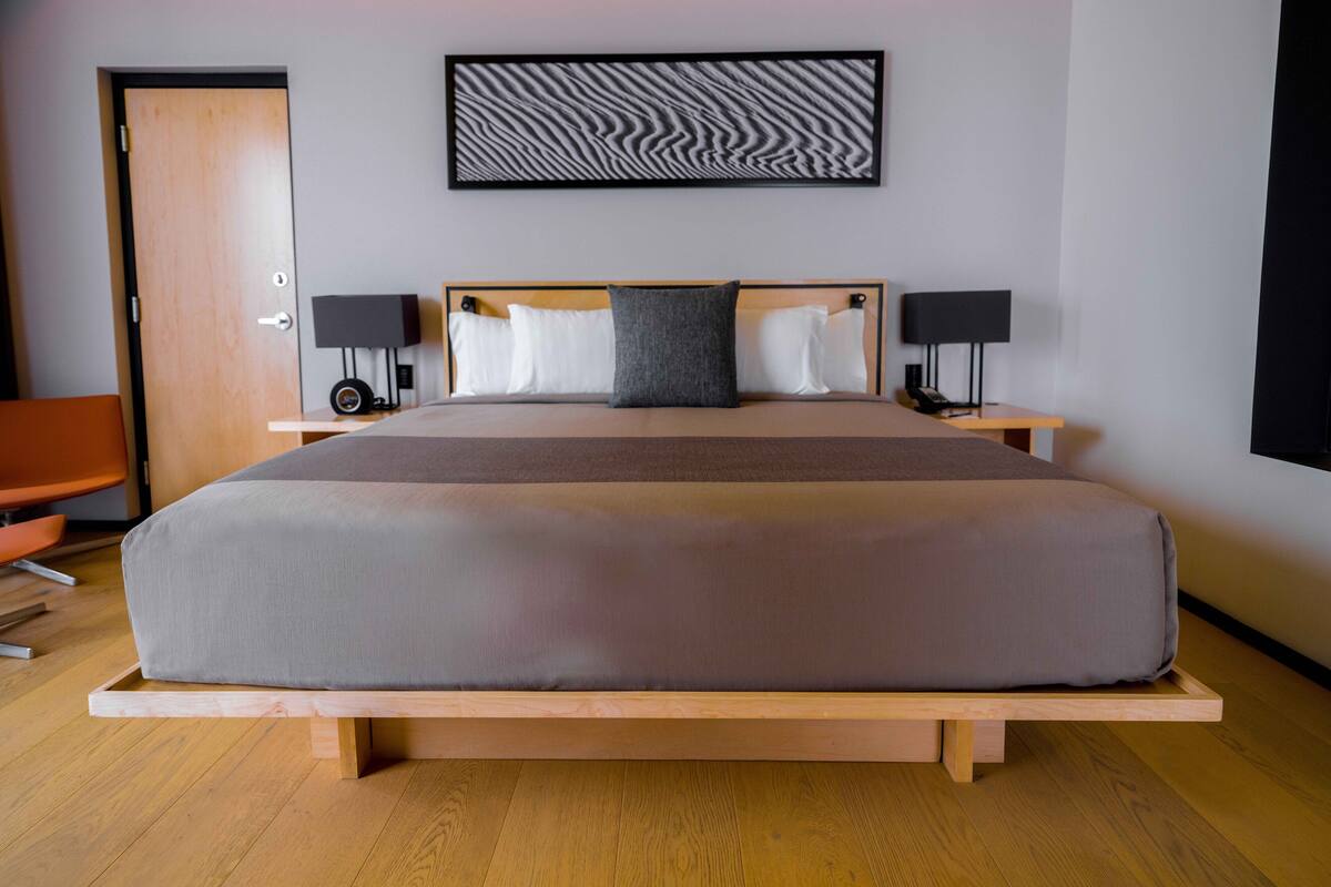 Una habitación de hotel moderna con una cama grande, dos mesitas de noche con lámparas, un banco de madera, una puerta y una obra de arte colgada en la pared.