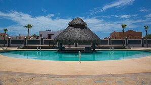 A swimming pool with a thatched-roof structure in the center, surrounded by palm trees and buildings. - Linda Vista 106 (2Bedroom-2Bathroom) By FMI Rentals (Puerto Peñasco)