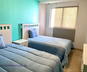 Una habitación con dos camas, una ventana con persianas, una mesita de noche y un aparador.