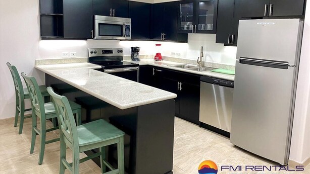 A modern kitchen with dark cabinets, a stainless steel refrigerator, and a central island with chairs. - Linda Vista 106 (2Bedroom-2Bathroom) By FMI Rentals (Puerto Peñasco)