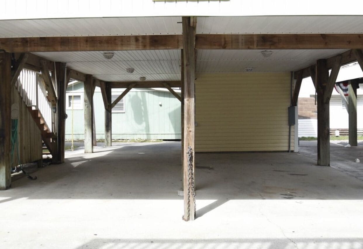 An empty covered parking area with concrete floor and wooden beams, featuring a yellow garage door and a staircase on the left.
