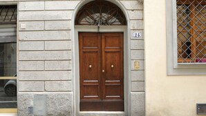 A double wooden door with a metal arch above it, a number '24' on the wall to the right, and a window with metal bars to the right of the door.
