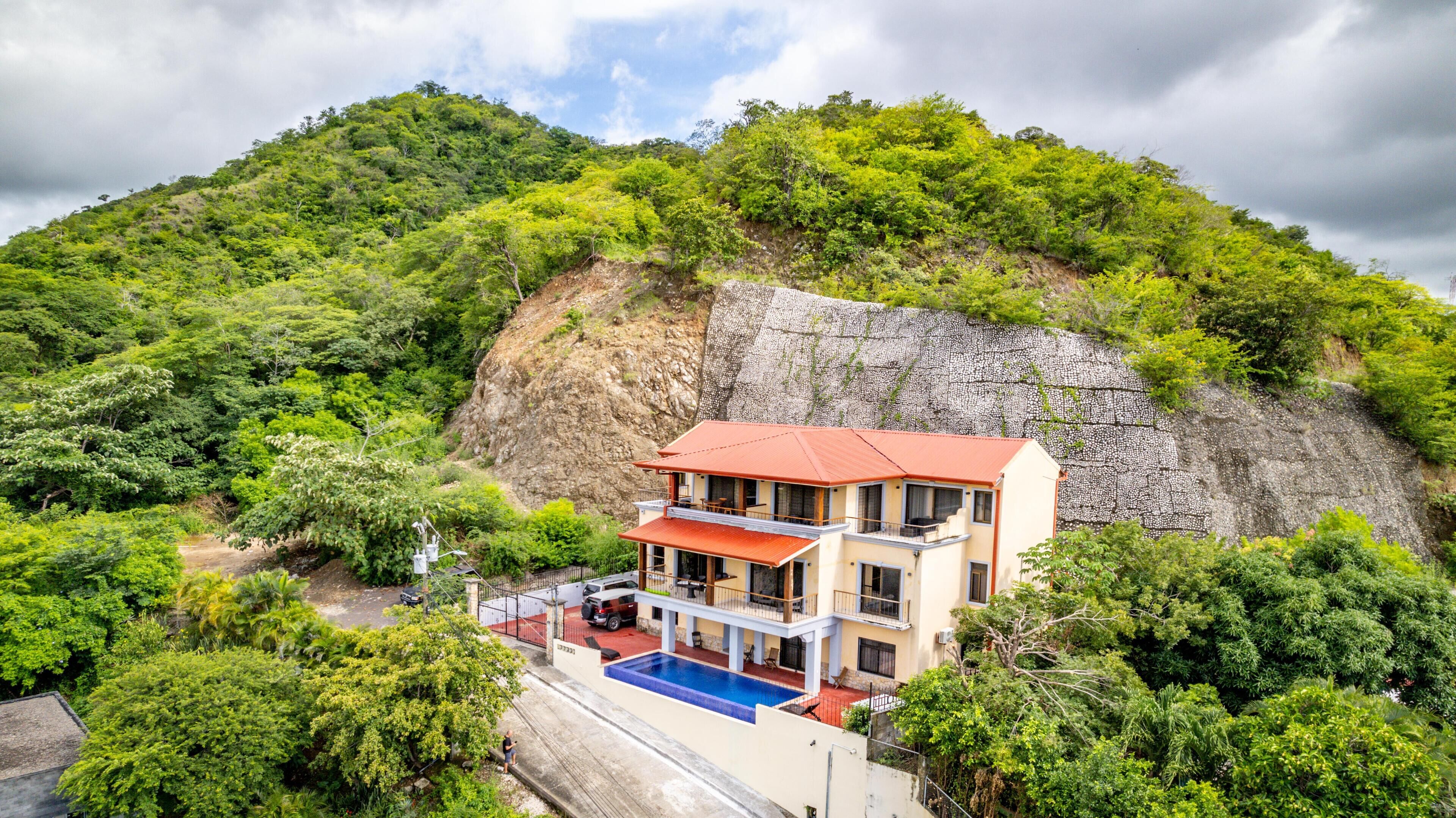 A two-story building with a red roof, a swimming pool, and a view of a lush green hillside.