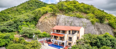 A two-story building with a red roof, a swimming pool, and a view of a lush green hillside.