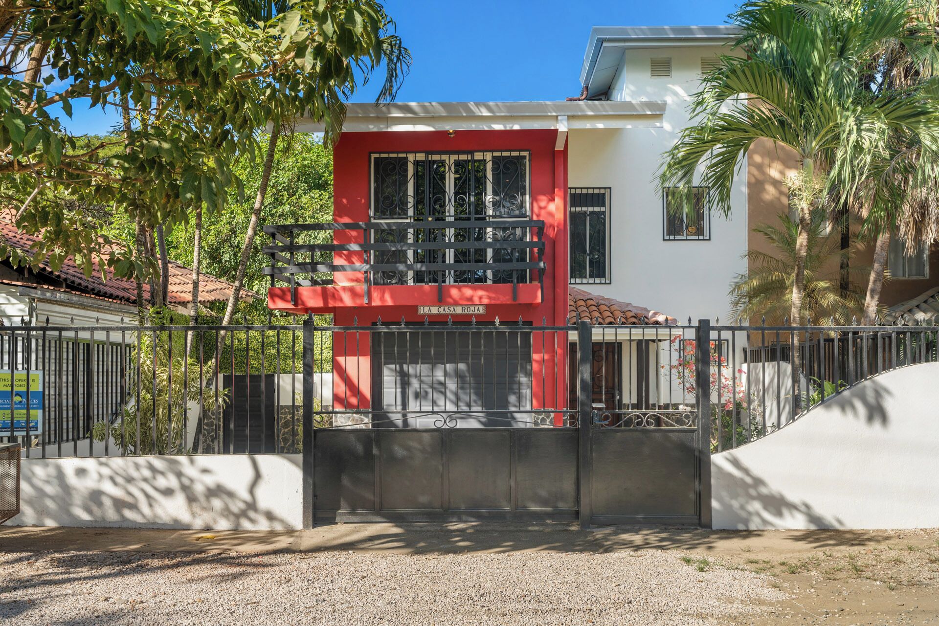 A gated entrance to a property with a red and white facade, a metal gate, and surrounding greenery.