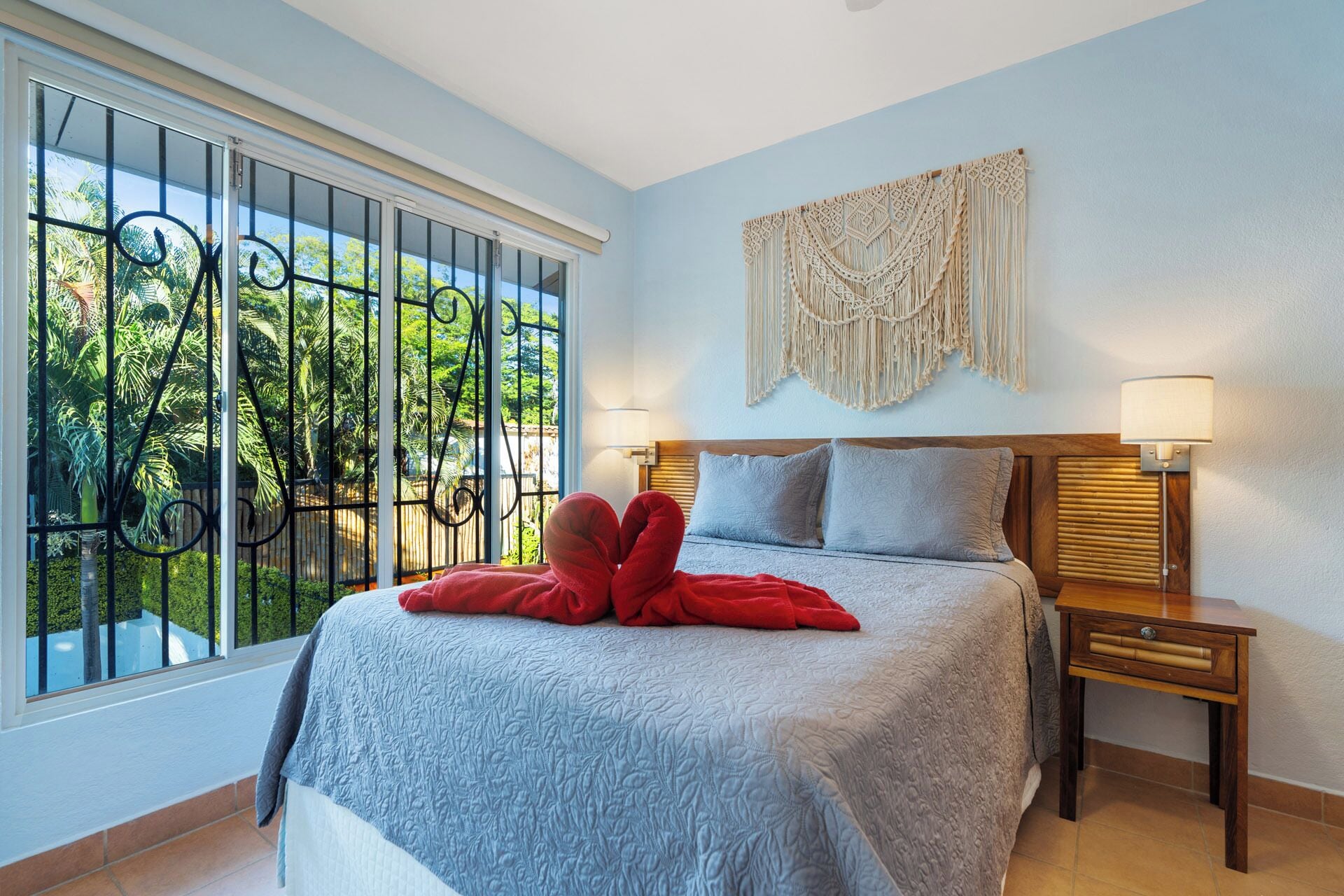 Ein Schlafzimmer mit einem Bett, zwei Nachttischen, einer Wanddekoration aus Makramee und Blick auf Grün hinter einem vergitterten Fenster.