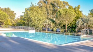 A rectangular swimming pool with a tiled deck, surrounded by trees and a metal fence. - Le Grand Parc by Interhome (Saint Aygulf)