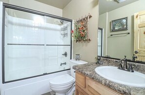 Een badkamer met een douche met glazen wand, een wit toilet, een wastafel met granieten blad, houten kastjes en een decoratief wandobject.