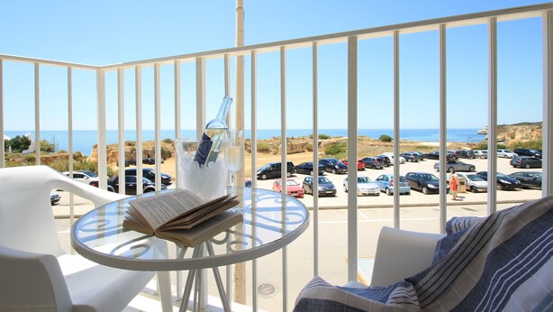 Un balcone con un tavolino in vetro, una sedia e un libro. Si intravedono auto parcheggiate e una spiaggia sullo sfondo.