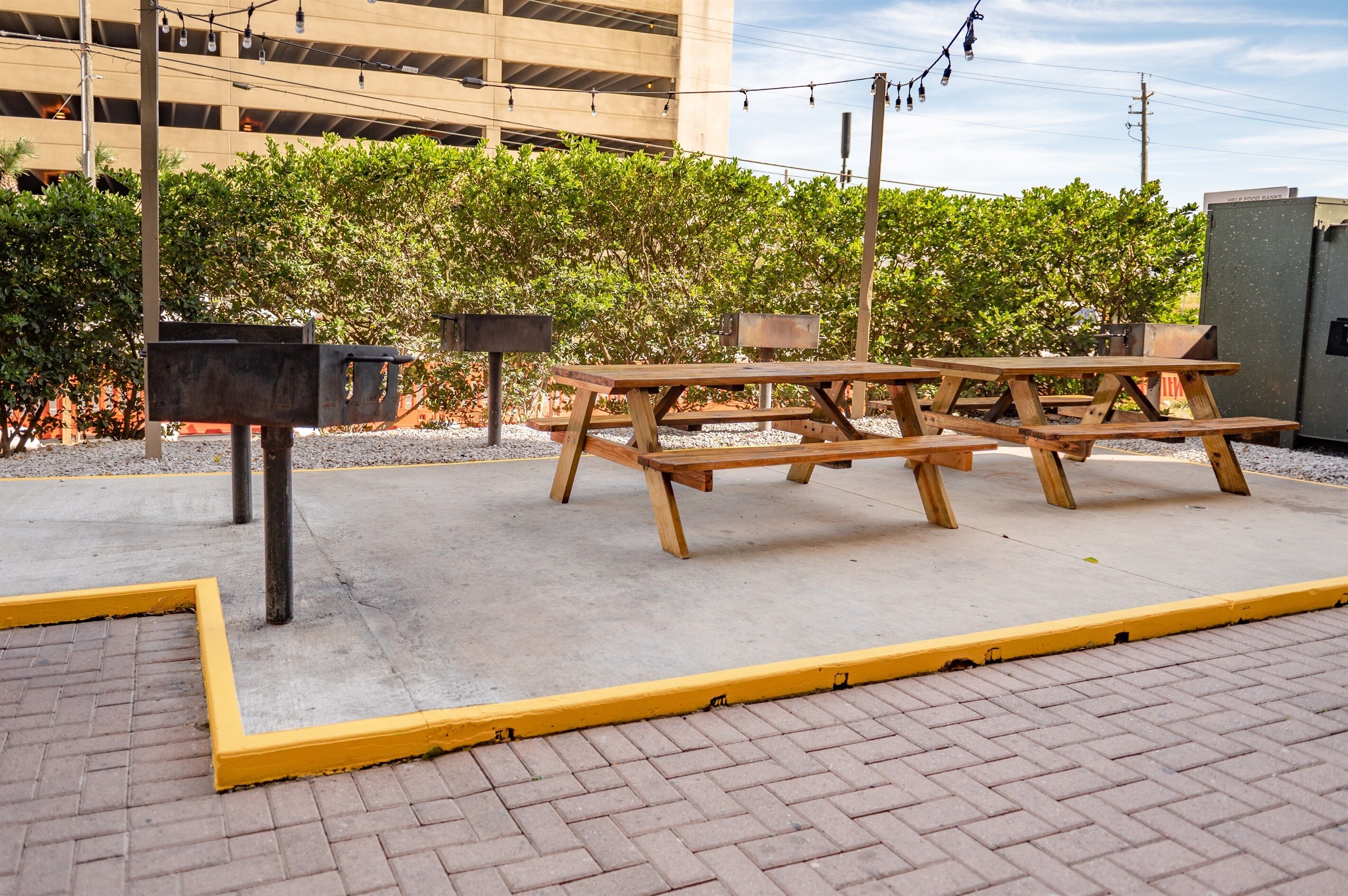 An outdoor picnic area with wooden benches and trash bins, surrounded by a hedge and a parking garage in the background.