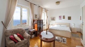 A bedroom with a bed, a wooden table, a sofa, a cabinet, and a view of the sea.