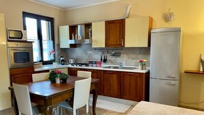A compact kitchen with wooden cabinets, a built-in oven, a refrigerator, and a dining area with a table and chairs. - Bnbook - La Corte di Gallarate (Gallarate)