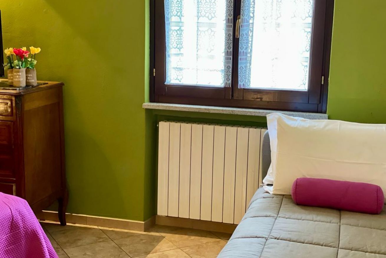 Una camera da letto con una parete verde, una finestra con tende in pizzo, un termosifone, un letto con cuscino bianco e rosa e un comò in legno con un vaso di fiori.