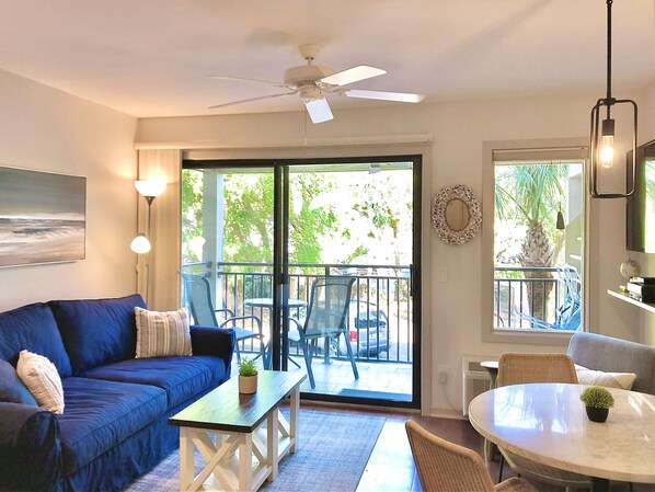 A living room with a blue sofa, a small table, a ceiling fan, and a balcony with chairs and a table.