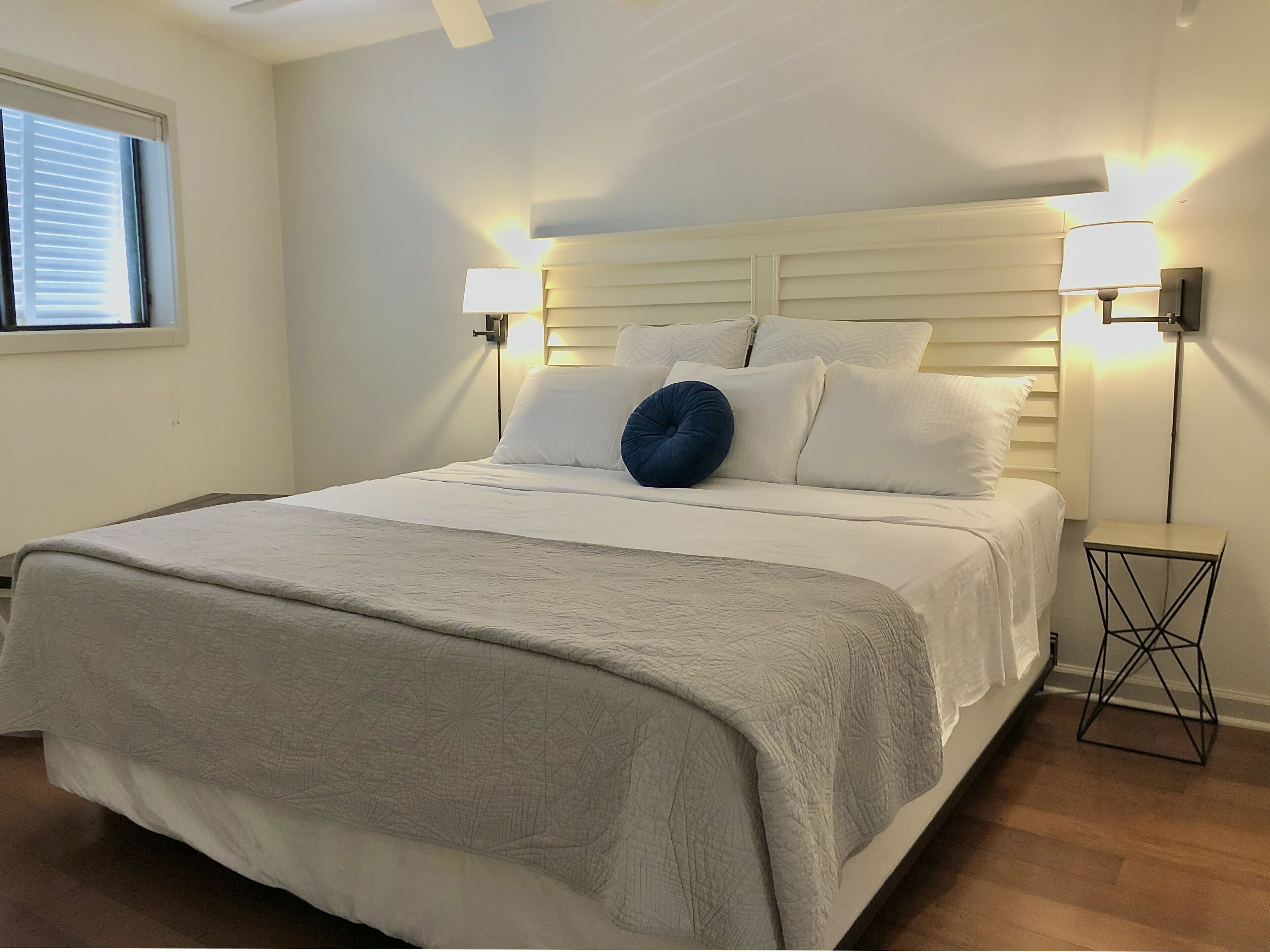 Una cama bien hecha con cabecera blanca, dos lámparas de noche y un almohadón azul.