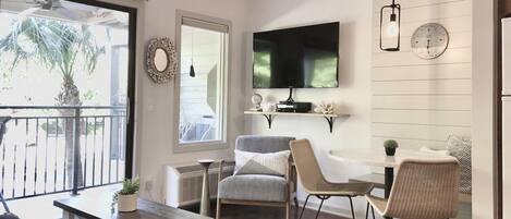 A modern living room with a flat-screen TV, comfortable seating, a dining area, and a balcony with a view.