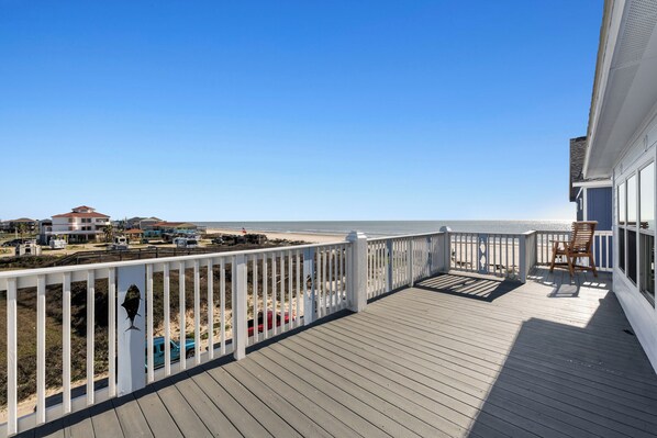 Una terraza de madera con barandilla blanca, con vista a una playa y edificios al fondo.