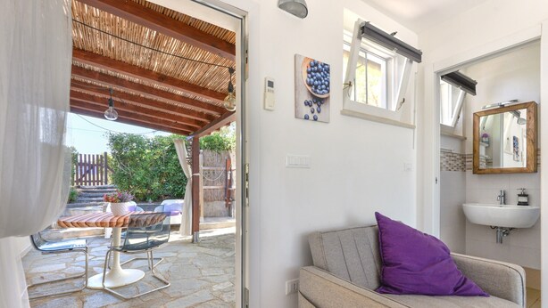 Una habitación con suelo de piedra, un sofá con cojín púrpura, techo de madera, lavamanos blanco y ventana con marco de madera.