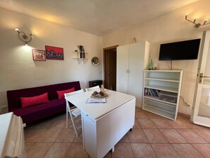 A compact living space with a white table, a purple sofa, a white cabinet, and a TV mounted on the wall. - Holiday Home "Villetta Lampone" with Air Conditioning, Garden and Terrace (CAPOLIVERI - LACONA)