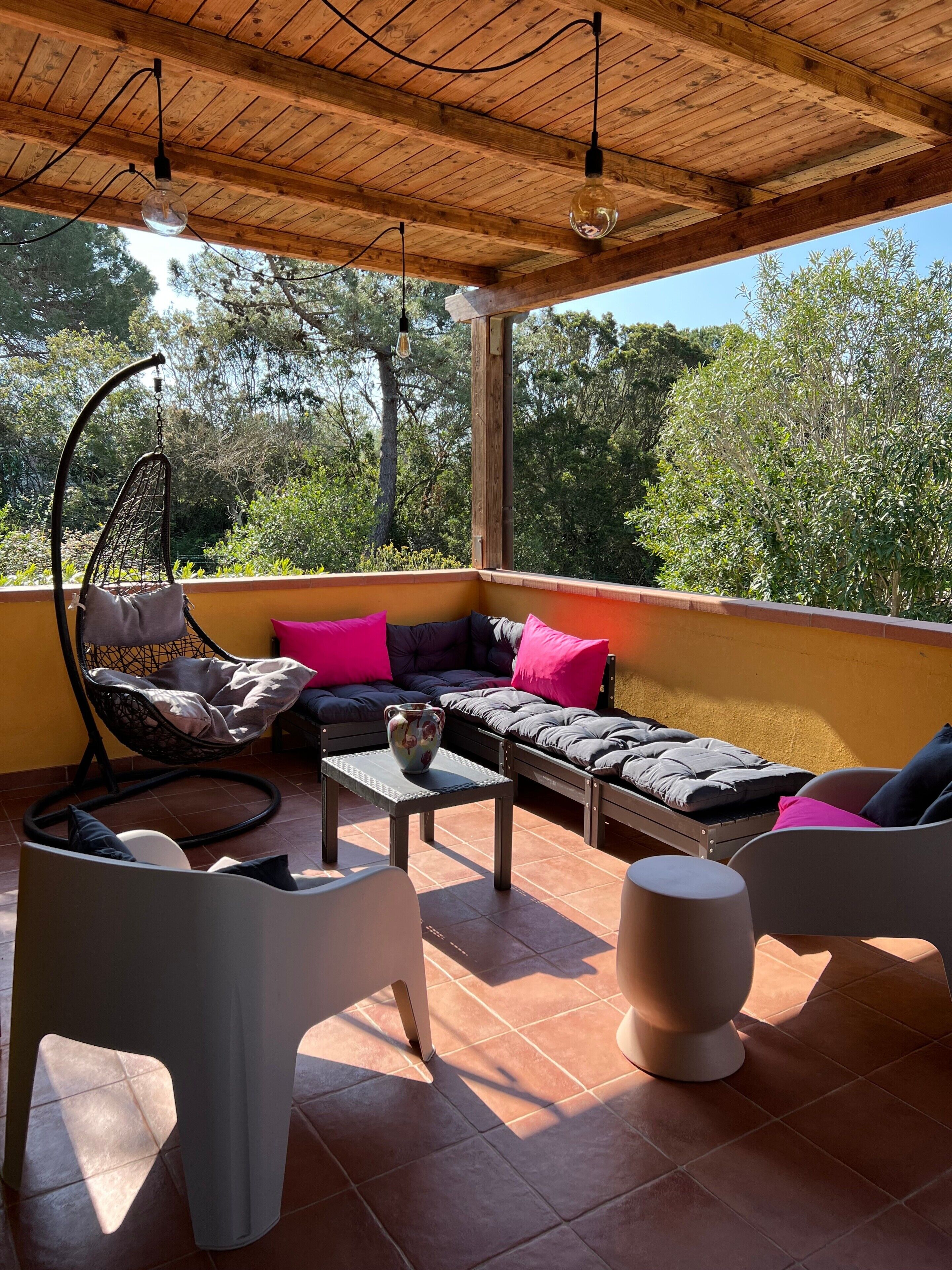 Un'area salotto esterna riparata, con tetto in legno, lampadario e vista sugli alberi.