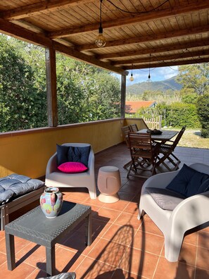 En overdækket terrasse med udendørs møbler, et bord og udsigt til grønt og bakker.