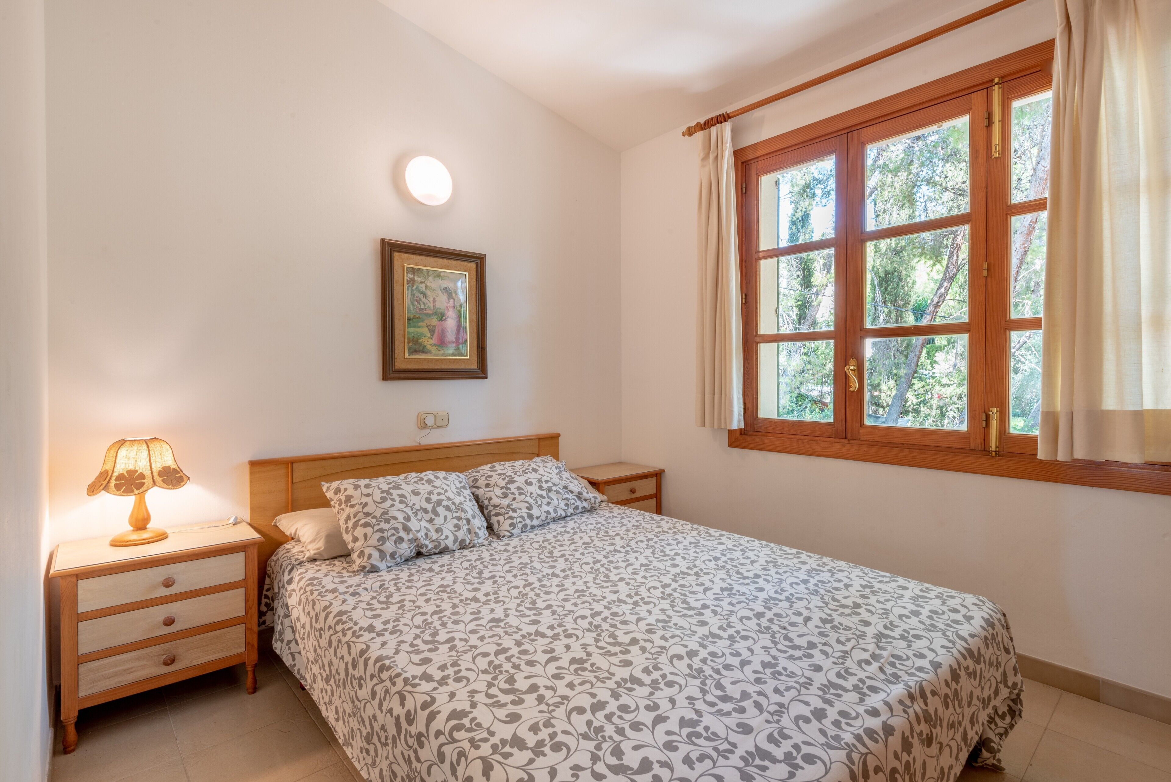 Ein Schlafzimmer mit einem Bett, einem Nachttisch mit Lampe, einem gerahmten Bild an der Wand und einem Fenster mit Vorhängen.
