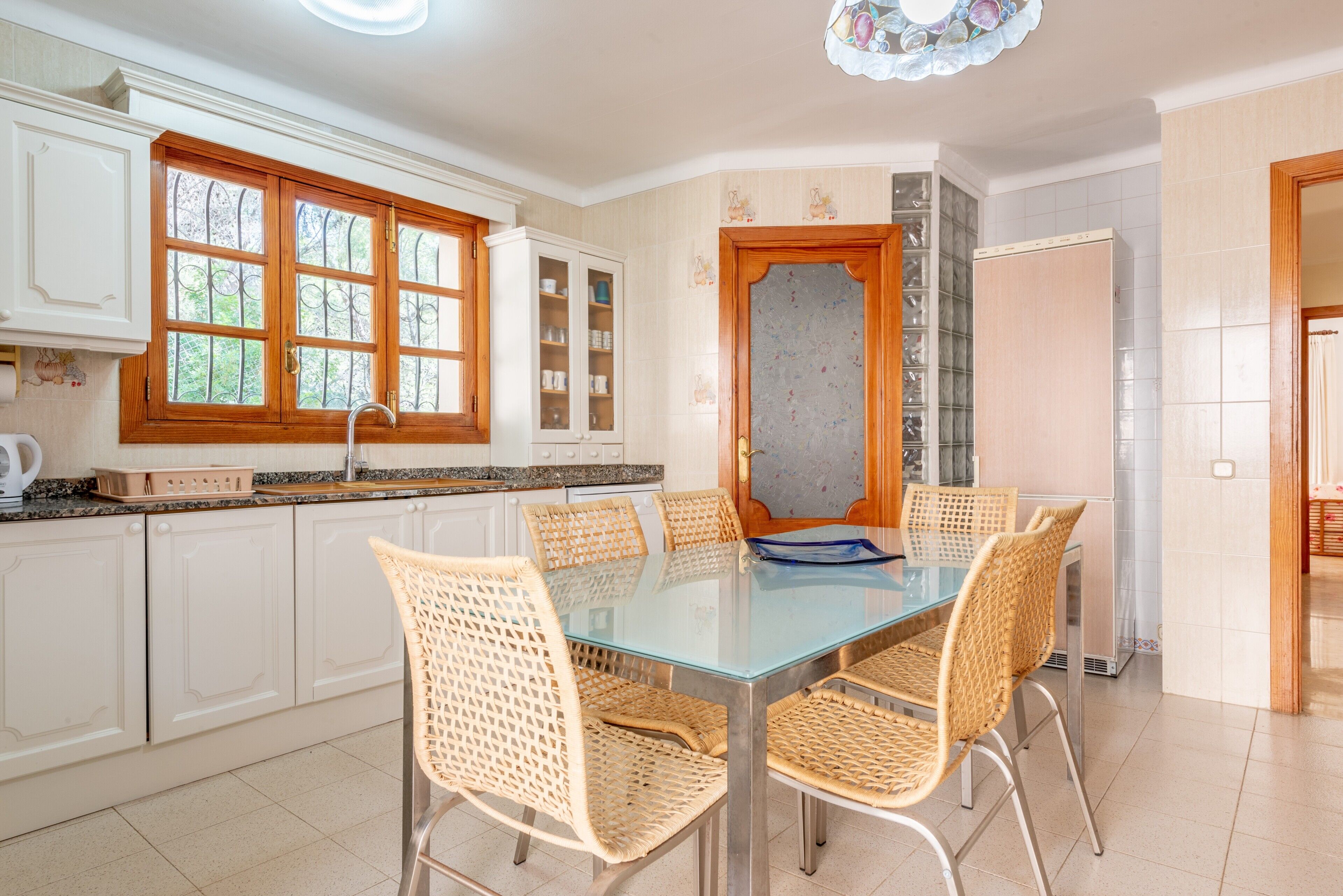 Una cocina con armarios blancos, una mesa de comedor de superficie de vidrio, sillas de mimbre y una ventana con rejas.