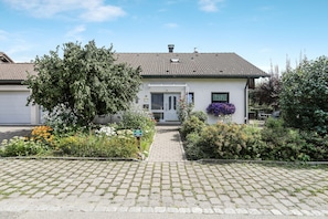 A single-story house with a paved driveway leading to a front door, surrounded by a garden with various plants and flowers.