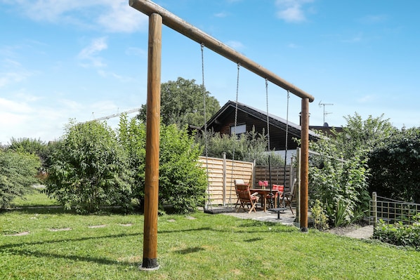 A wooden swing set in a grassy backyard with a wooden fence, a dining table with chairs, and a building in the background.