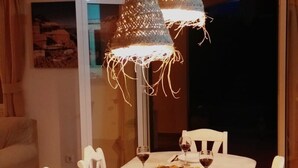 Un coin repas avec une table dressée pour deux personnes, deux lampes suspendues, des chaises blanches et une bouteille de vin.