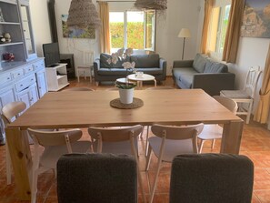 A dining area with a wooden table and white chairs, a sofa, and a television in the background.