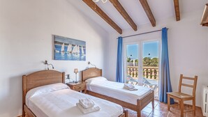 Une chambre à coucher avec deux lits en bois, une table de chevet, une chaise, un tableau représentant des voiliers et une vue sur la mer par une porte vitrée.