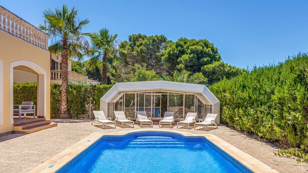 Une piscine avec des chaises longues, un espace de détente couvert et un bâtiment doté d’une entrée en arc de cercle.