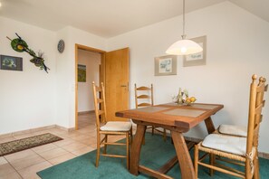 Un coin repas avec une table en bois, des chaises en osier, un luminaire suspendu et une porte donnant sur une autre pièce.