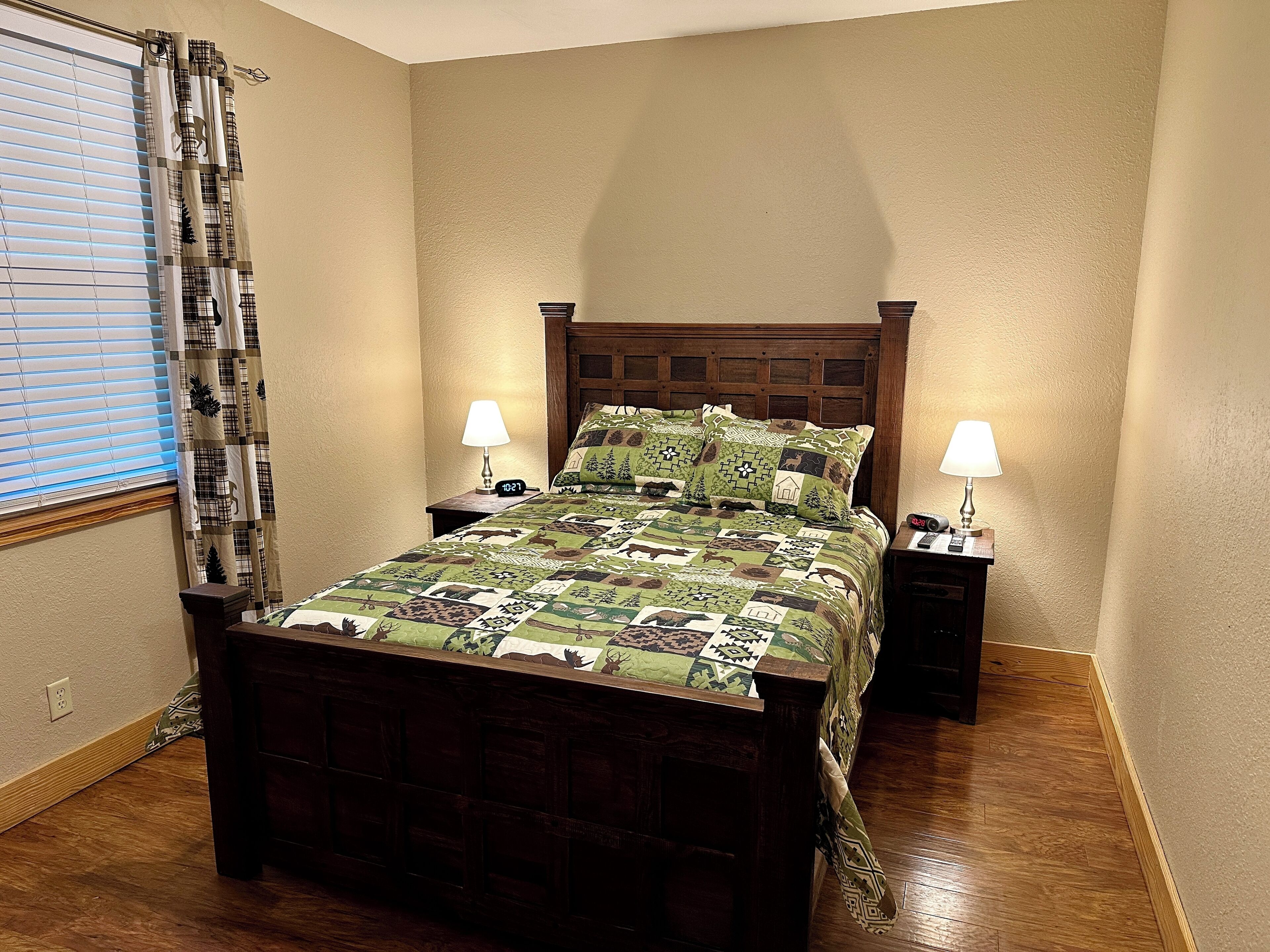 A bedroom with a wooden bed, two bedside tables with lamps, a clock, and a window with blinds.