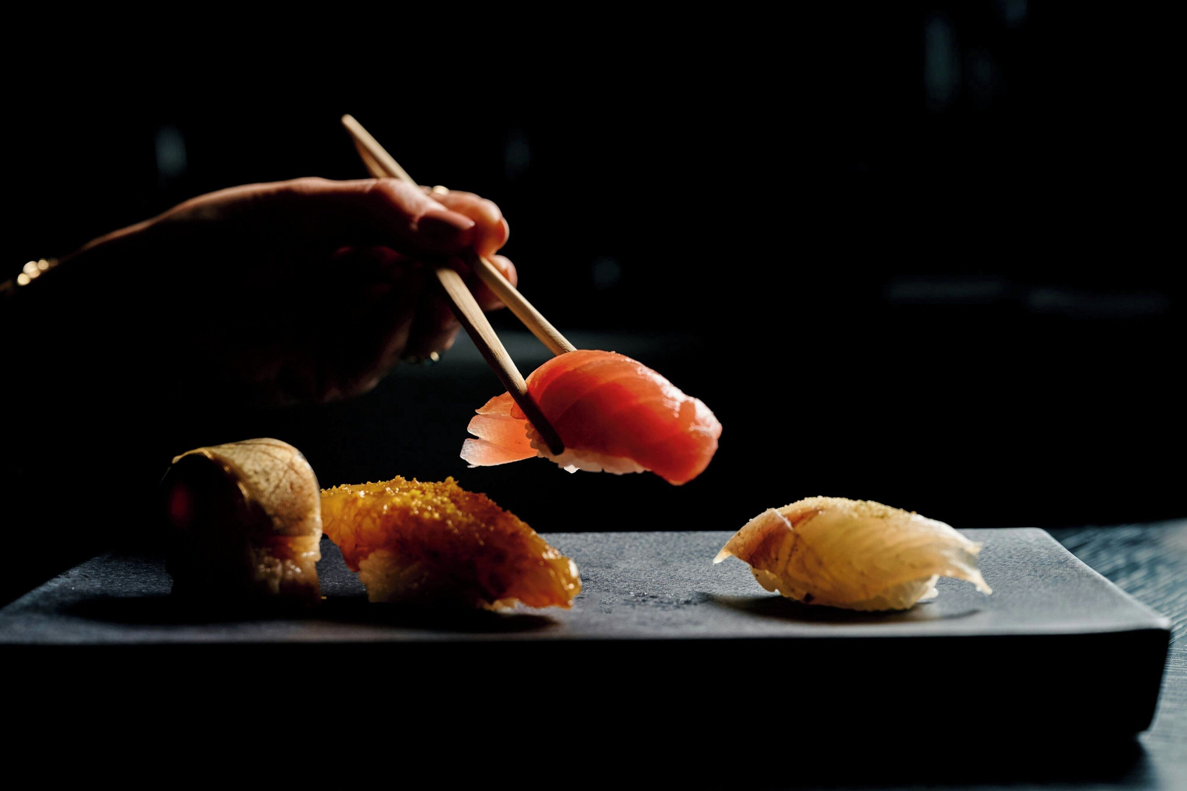 A hand holding chopsticks over sushi on a dark plate.