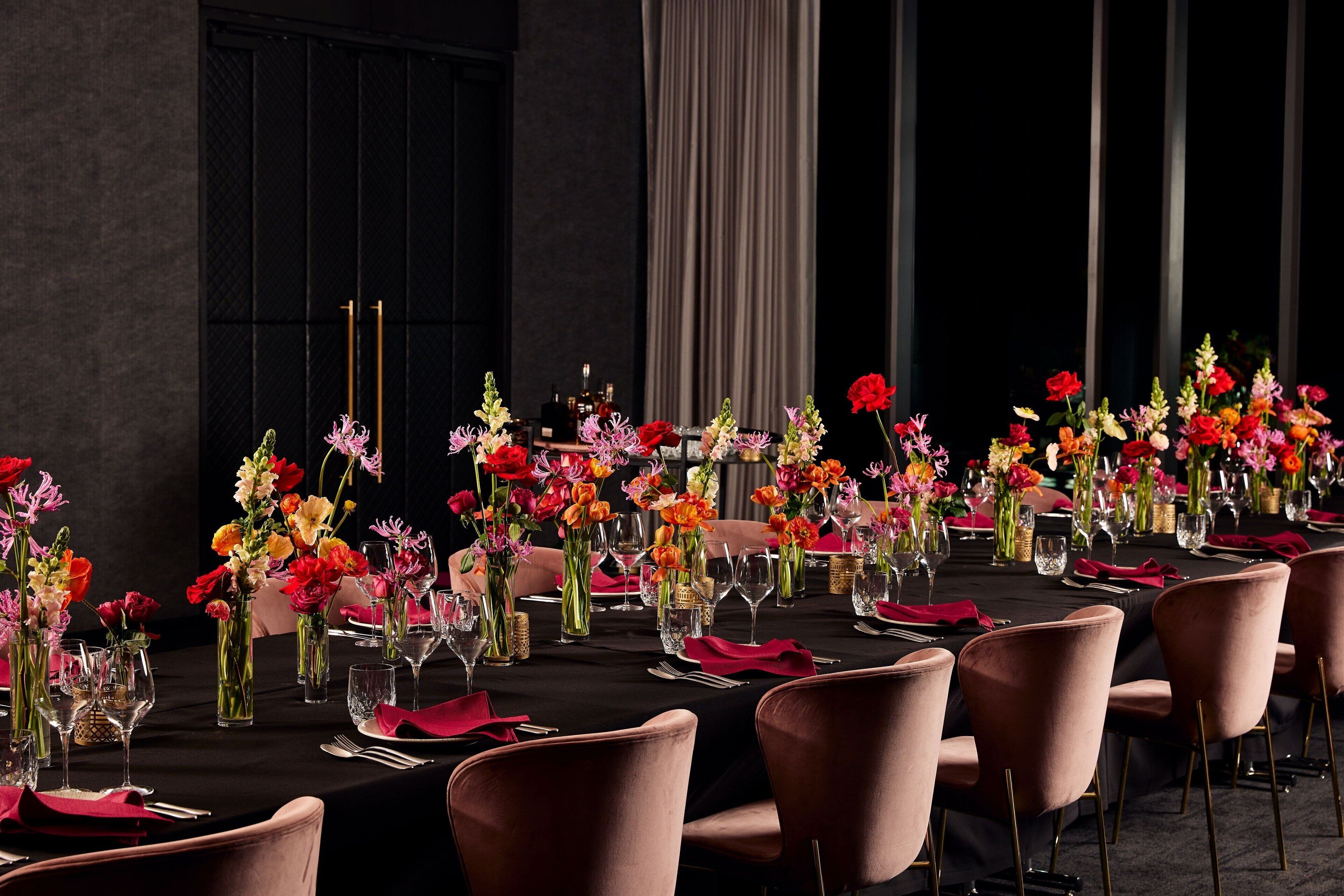 Una larga mesa de comedor con sillas rosas, decorada con flores coloridas en jarrones de vidrio y un mantel oscuro.