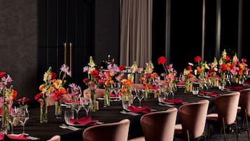 Una larga mesa de comedor con sillas rosas, decorada con flores coloridas en jarrones de vidrio y un mantel oscuro.