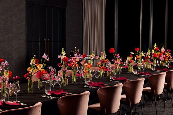 A long dining table set with pink chairs, adorned with colorful flowers in glass vases, and a dark tablecloth.