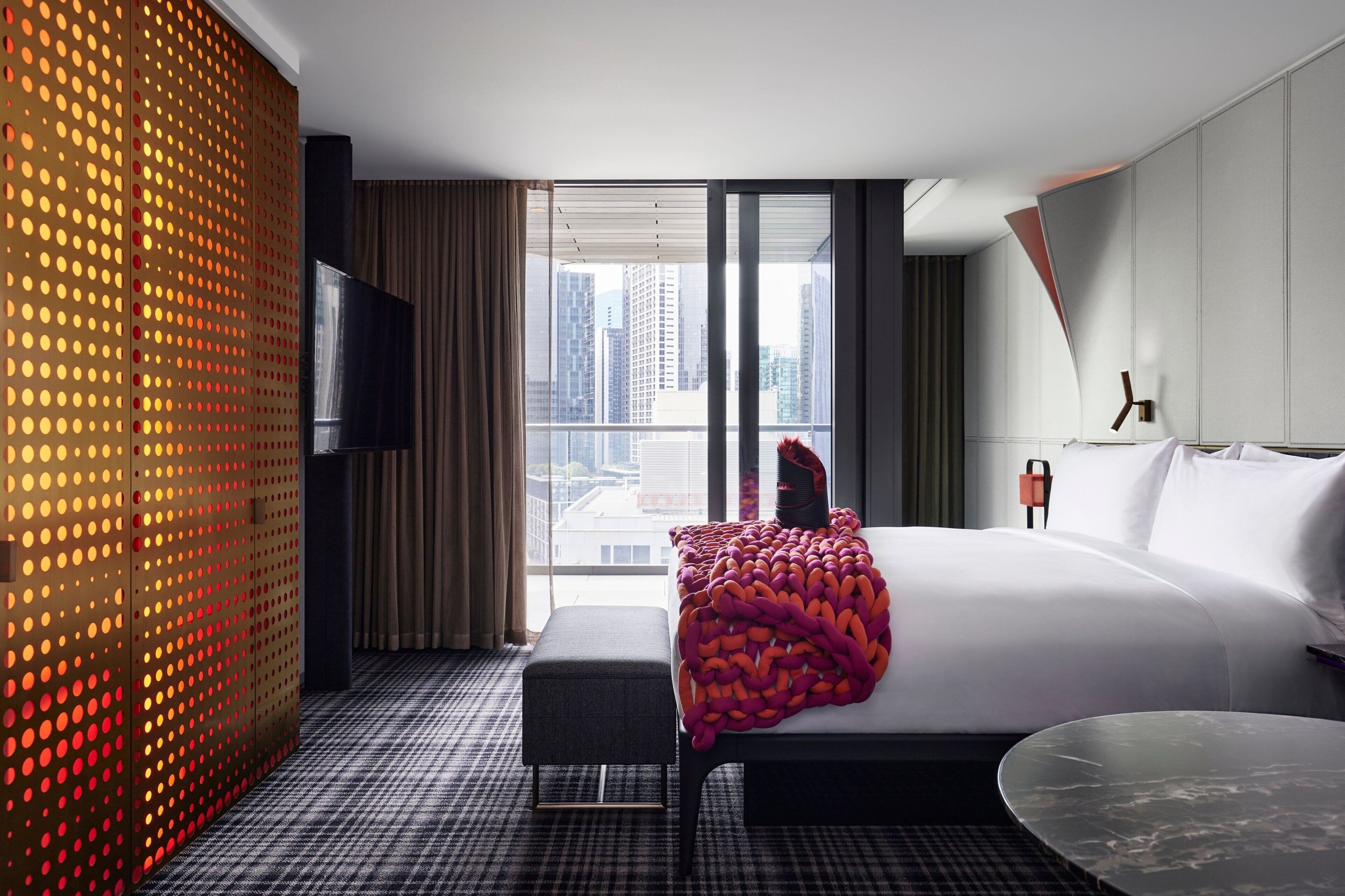 A modern hotel room with a large bed, a flat-screen TV, a city view, and a patterned floor.