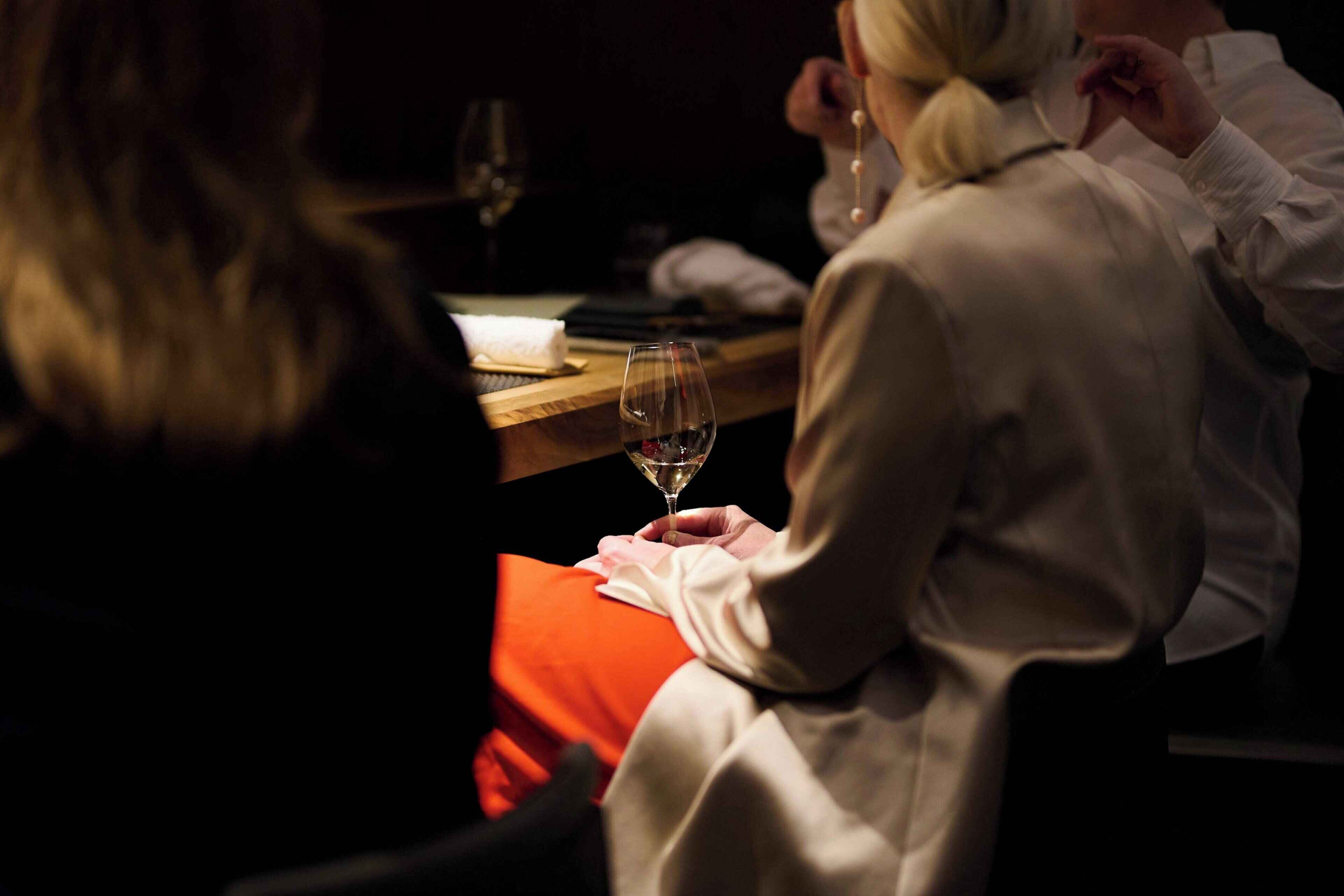 A person holding a wine glass, seated at a table with a wooden surface and a red cloth.