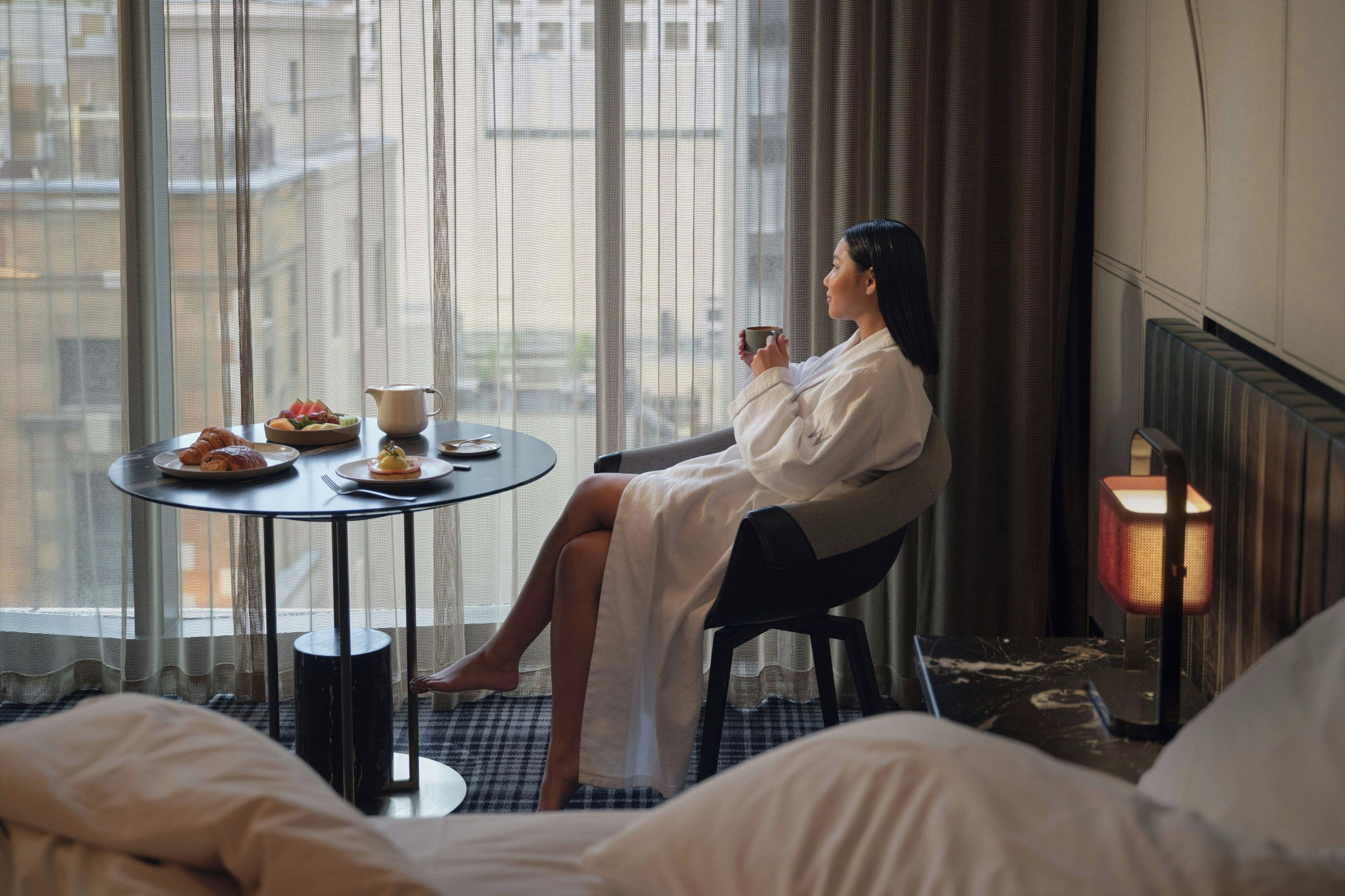 A hotel room with a bed, a round table with breakfast items, a person in a robe, and a window with curtains.