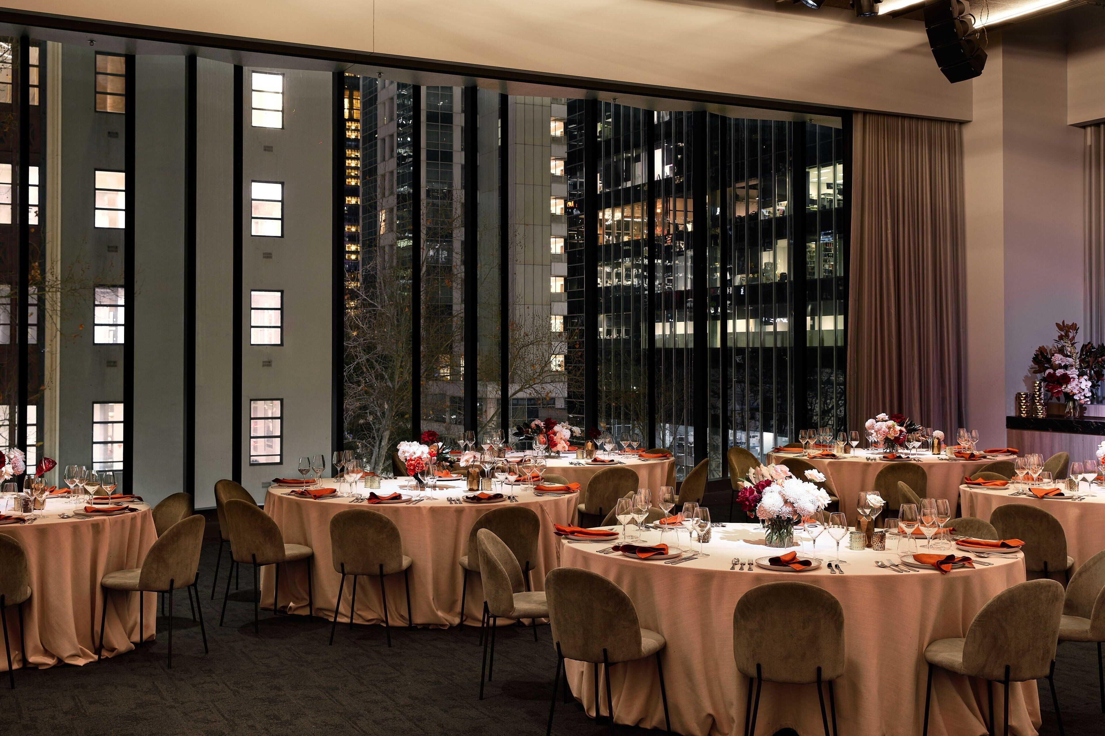 Une salle de banquet avec des tables rondes dressées pour le repas, de grandes fenêtres ornées de rideaux et une vue sur le paysage urbain illuminé la nuit.