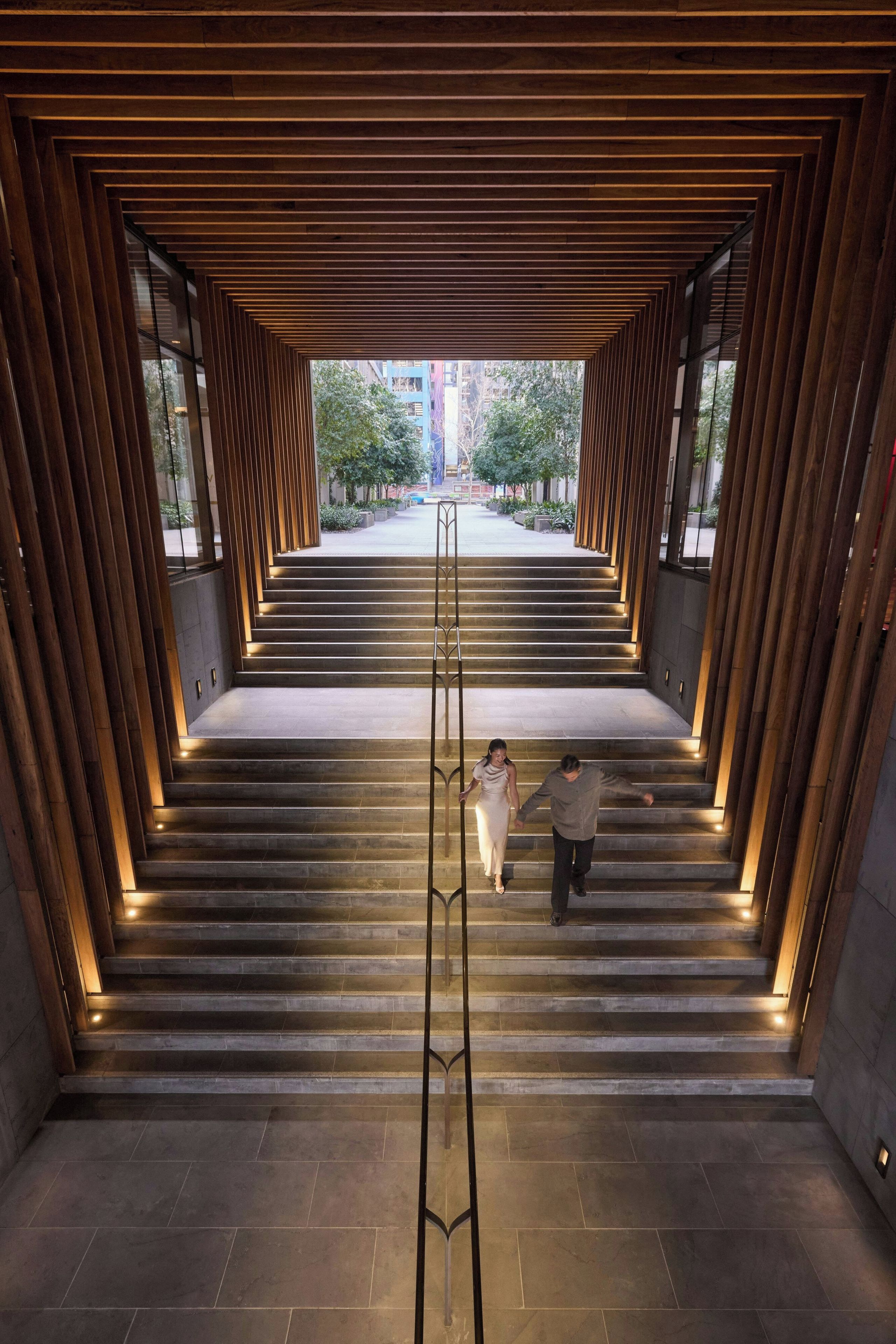 Eine moderne Treppe mit Holzverkleidung und Beleuchtung, die zu einem Außenbereich mit Bäumen und Gebäuden führt.