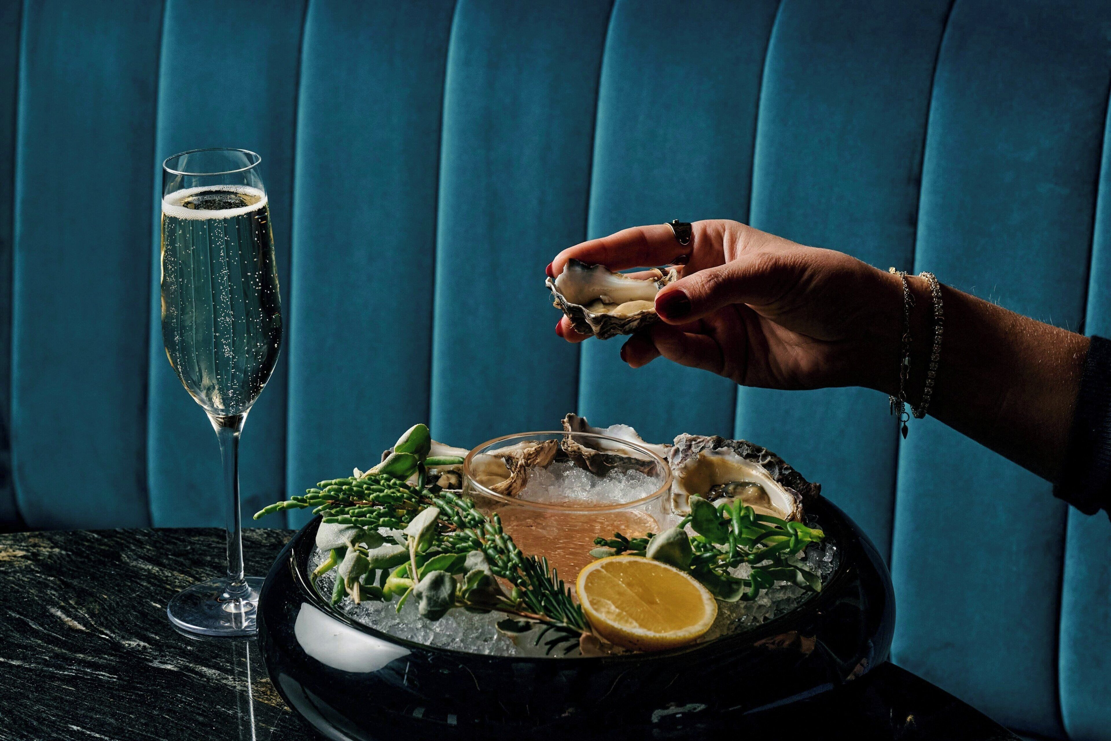 A hand holding an oyster with a glass of champagne and a bowl of oysters on a table.