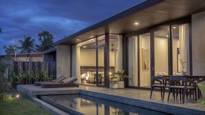 A modern outdoor pool area with a lounge chair, a dining table with chairs, and a well-lit interior space with a bed and a dining area. - Amber Kampot (Kampot)