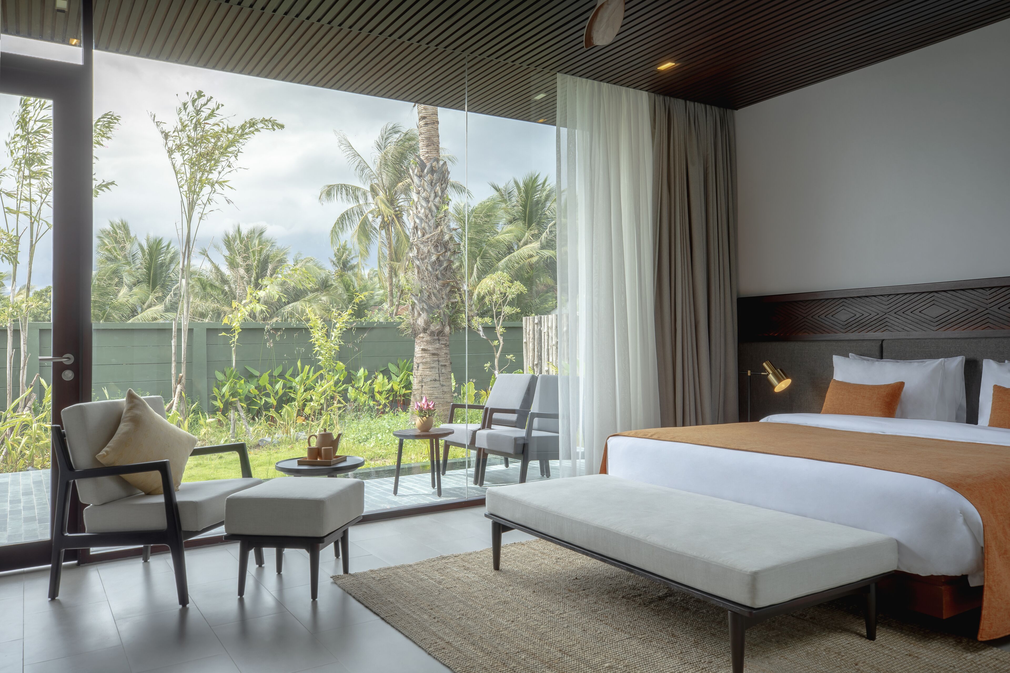 Habitación de hotel moderna con una cama grande, dos sillones, una mesita y vistas a palmeras y vegetación.