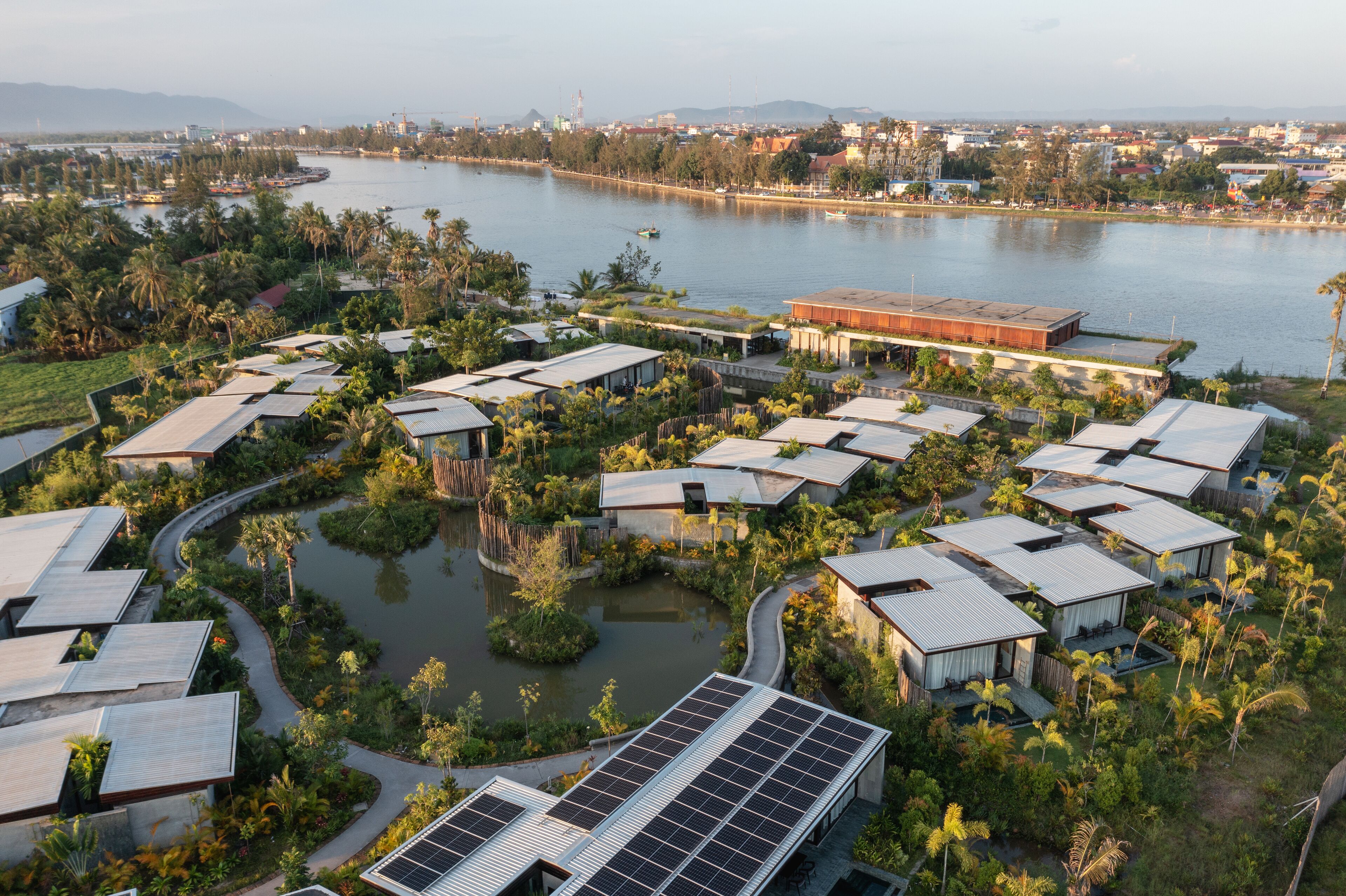A resort with multiple bungalow-style buildings, a central water feature, and a view of a river and distant cityscape.