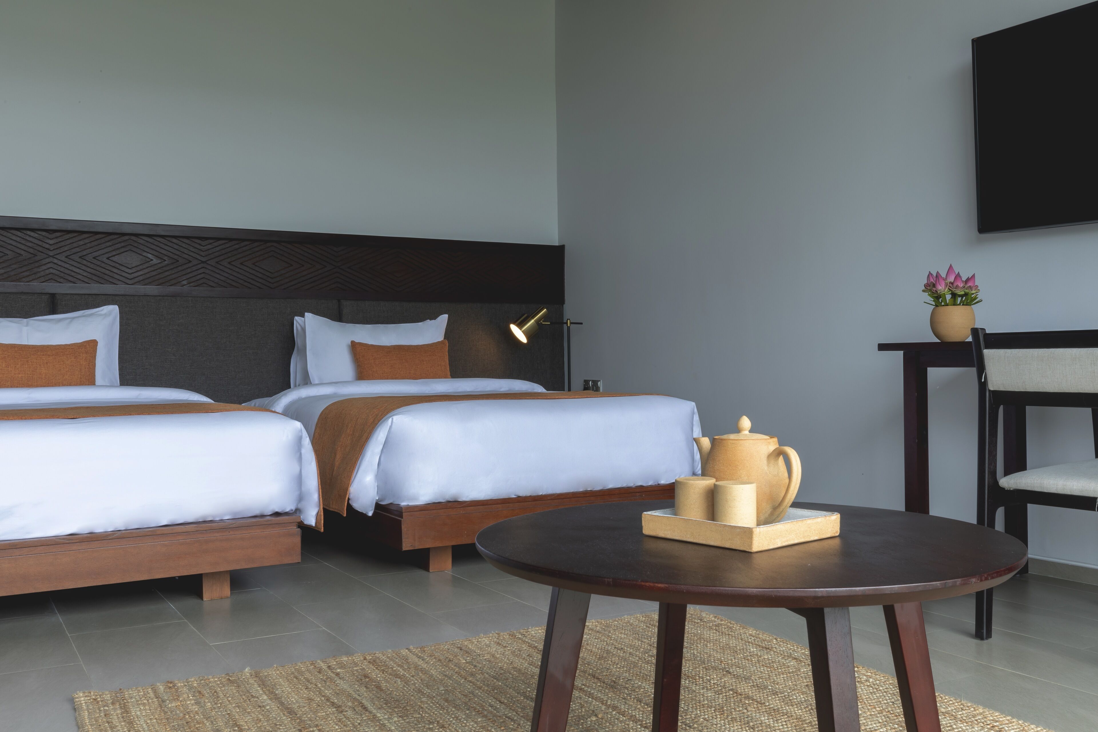 A hotel room with two beds, a round table, a chair, a TV, and a small potted plant.