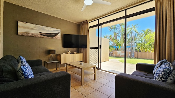 A living room with a flat-screen TV, two sofas, a coffee table, and a large sliding glass door leading to a garden. - Riverview Holiday Apartment 60 - Ground Floor -  Kalbarri WA (Kalbarri)